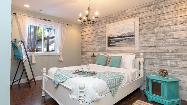 A bedroom with a wooden wall, a bed with a white headboard, a small teal nightstand, and a chandelier. -  2 Bedroom Suite @ Shawnigan Suite Bed and Breakfast (Shawnigan Lake)