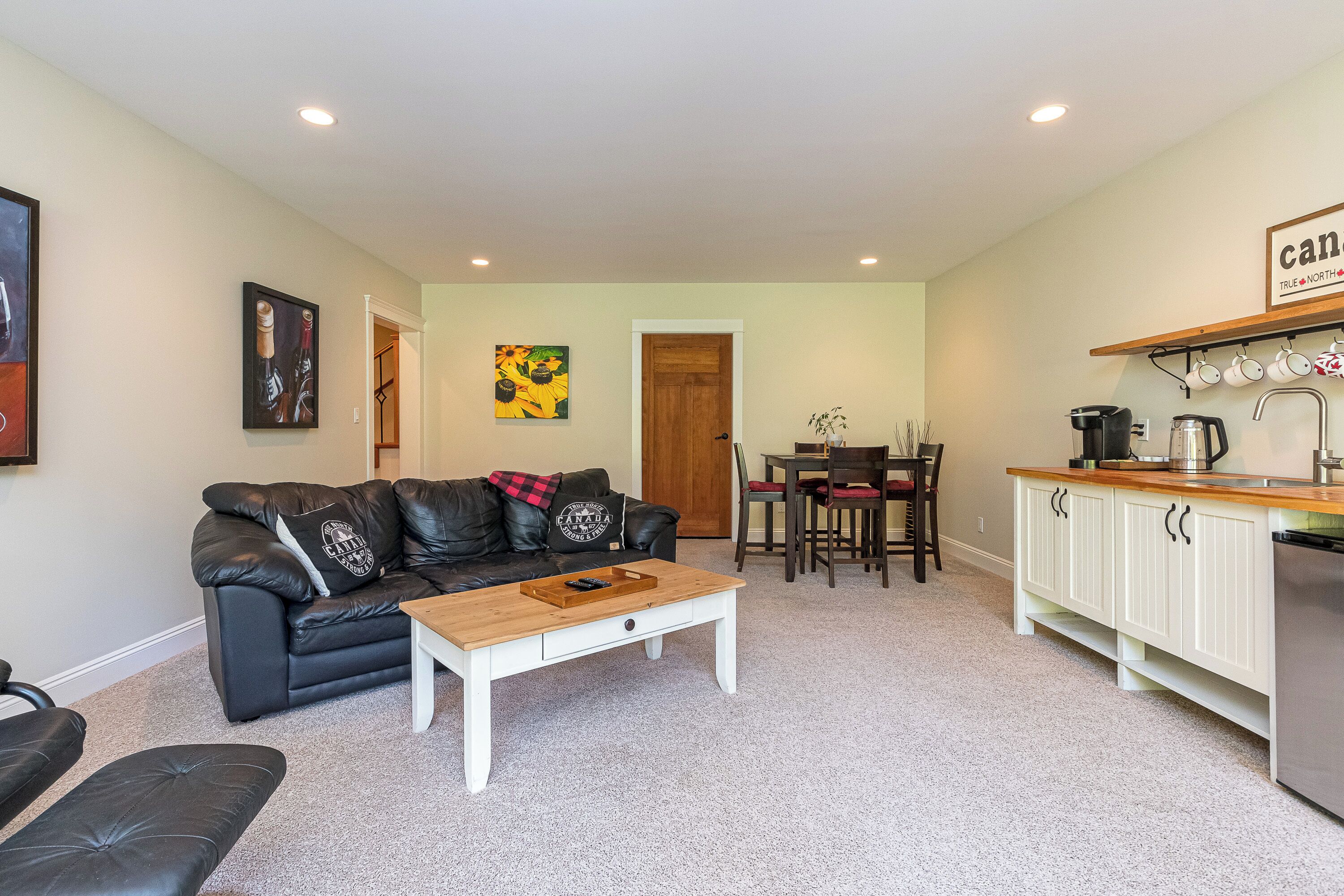 A compact living space with a black leather sofa, a wooden coffee table, a dining table with chairs, and a kitchen area with white cabinets and a countertop.