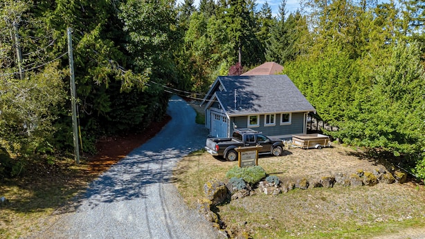 A two-story house with a gravel driveway, surrounded by dense forest.