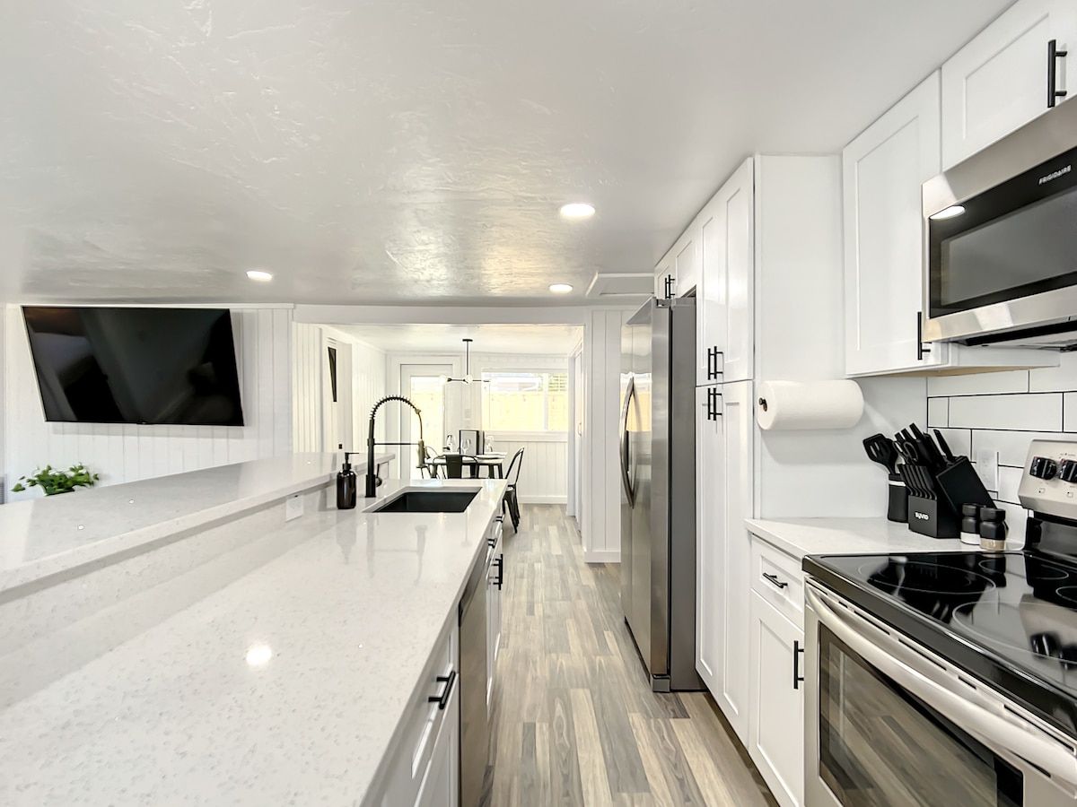A modern kitchen with white cabinetry, a stainless steel refrigerator, a built-in oven, and a flat-screen TV mounted on the wall.