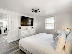 A bedroom with a large bed, a TV mounted on the wall, a dresser, and a window with blinds.