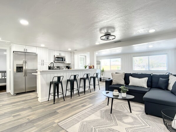A modern kitchen with a central island, bar stools, a dark sofa, and a patterned rug.