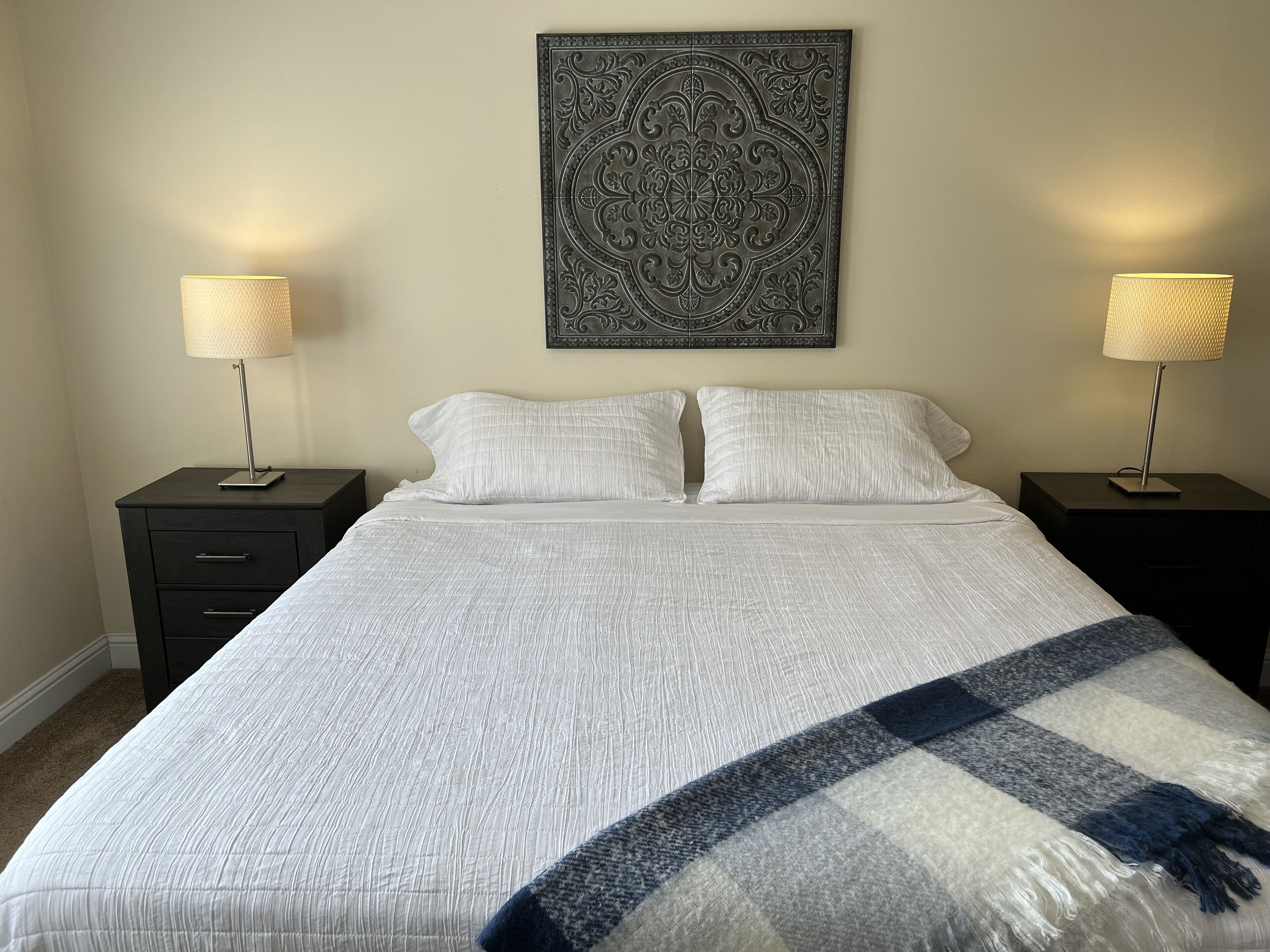 Una cama bien hecha con edredón blanco y dos almohadas, flanqueada por dos mesitas de noche con lámparas, y un adorno decorativo en la pared superior.