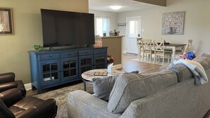A living room with a grey sofa, a blue cabinet with glass doors, a flat-screen TV, and a dining area with a wooden table and chairs. - Plum Peachey - Bay St Louis (Bay St. Louis)