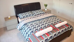 Een netjes opgemaakt bed met beddengoed met een patroon, een klein nachtkastje met een plantje, en een deur op de achtergrond.