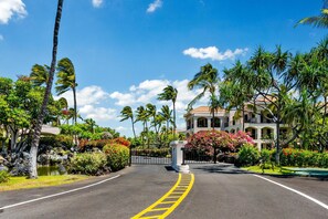 Une route menant à un bâtiment dont l’entrée est protégée par un portail, entouré de palmiers et d’une végétation tropicale.