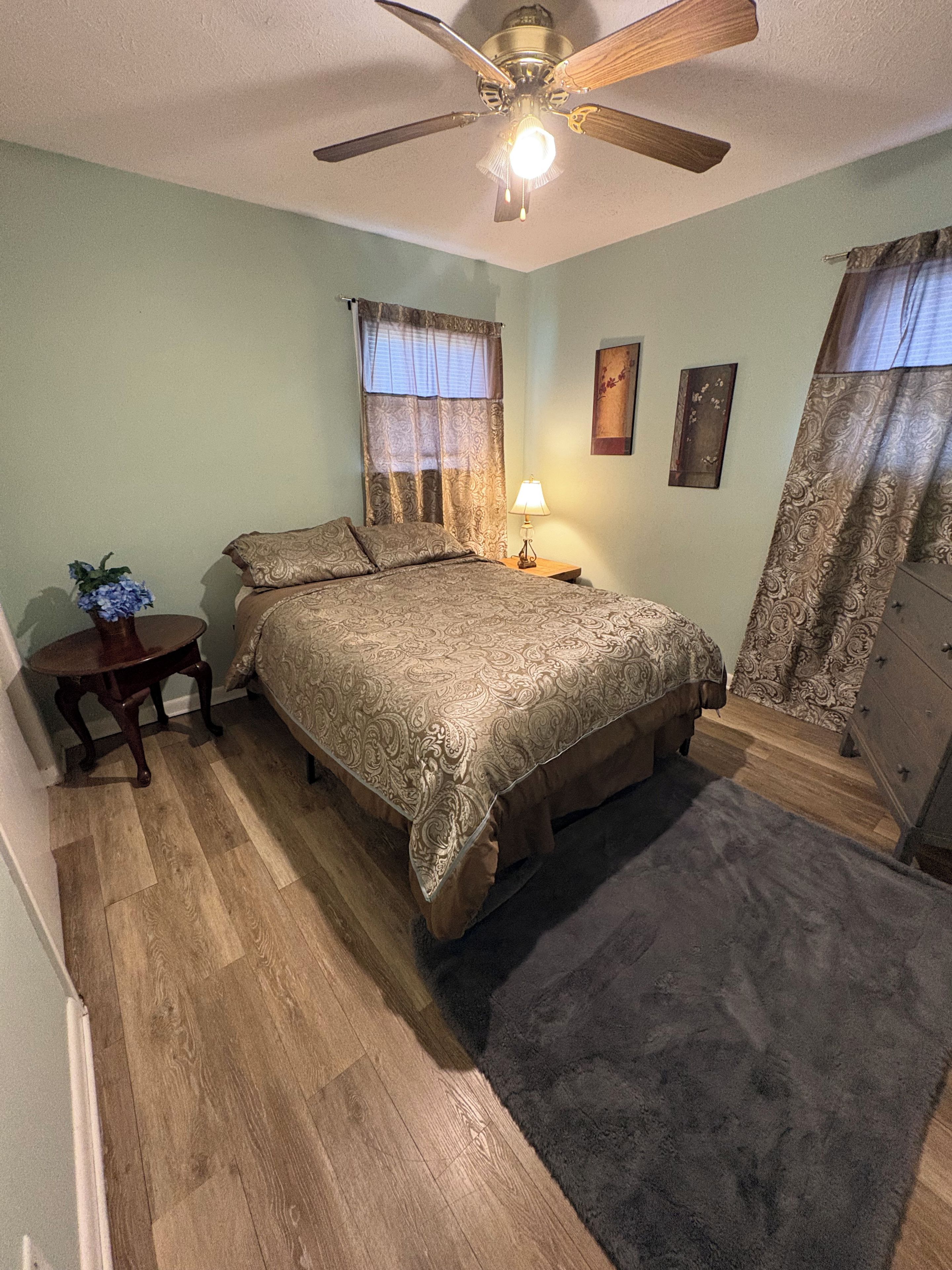 A bedroom with a bed, a nightstand, a lamp, a ceiling fan, and a dresser.