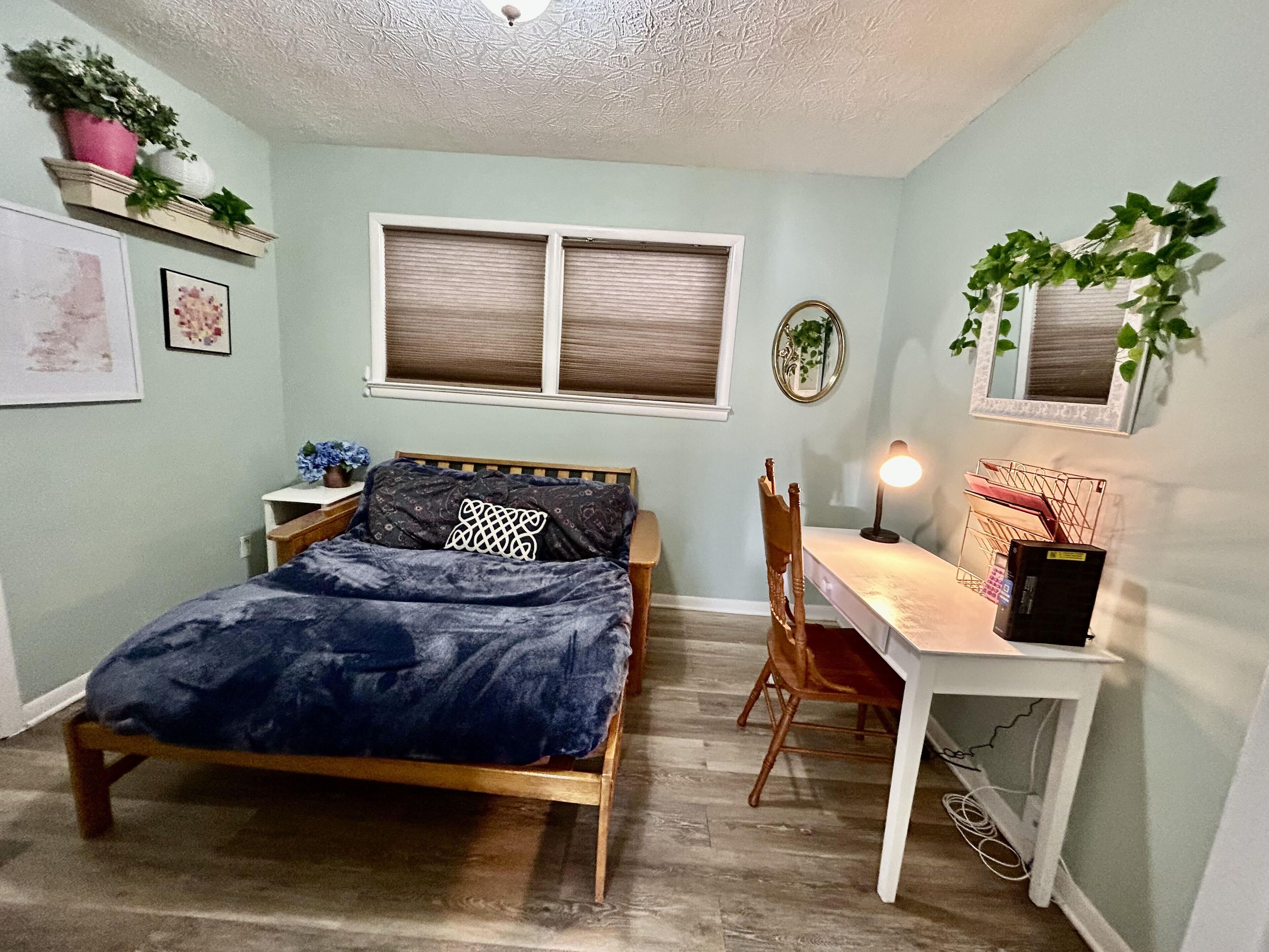 A bedroom with a bed, a desk, a chair, a window with blinds, a ceiling fan, and wall decorations including plants and framed pictures.
