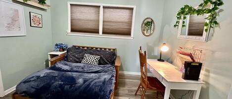 A bedroom with a bed, a desk, a chair, a window with blinds, a ceiling fan, and wall decorations including plants and framed pictures.