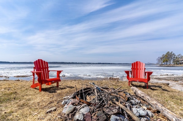 Zwei rote Adirondack-Stühle vor einem zugefrorenen See unter blauem Himmel.