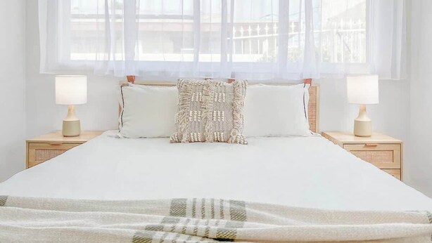 A neatly made bed with a white bedspread and two pillows, flanked by bedside tables with lamps, in a room with large windows and sheer curtains.