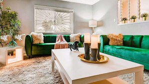 A living room with a green sofa, a white coffee table, a decorative mirror, and a window with a geometric design.