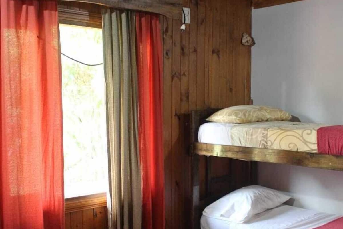 Une chambre avec un lit superposé, une couette rouge, des murs en bois et une fenêtre avec des rideaux.