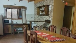 A dining area with a wooden table and chairs, a kitchen in the background, and a stone wall. - Raices Pampeanas (Puerto Iguazú)