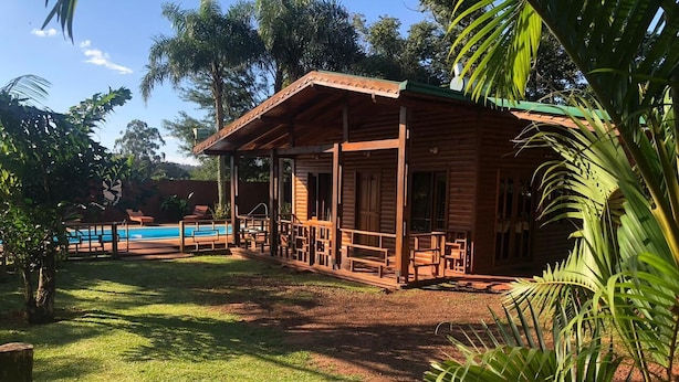 A wooden cabin with a porch, surrounded by lush greenery and a pool in the background. - Raices Pampeanas (Puerto Iguazú)