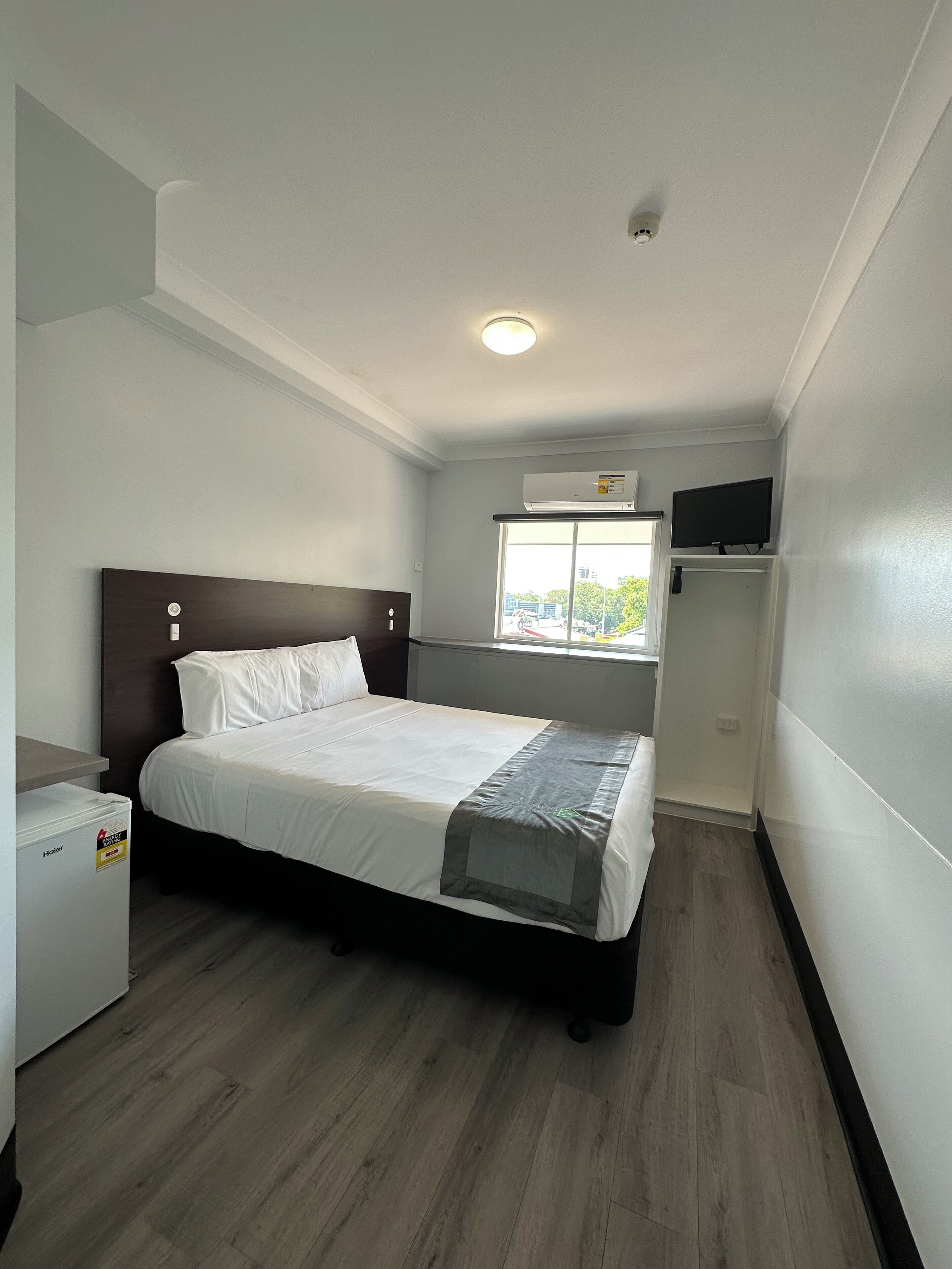 A small, neatly arranged bedroom with a bed, a small heater, a window, and a television on a stand.