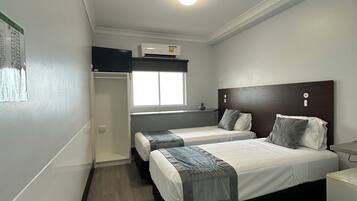 A compact hotel room with two beds, a small desk, a window, and a ceiling-mounted air conditioner.