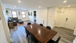 A spacious living area with a wooden dining table, a sofa, and a coffee table. There is a kitchen area with white cabinets and a door. - Downtown 3 Bed Loft Apartment (Delaware)