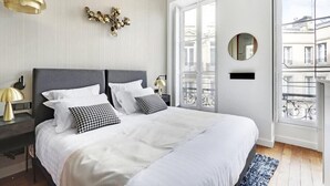 A bedroom with a large bed, a bedside table, a lamp, a wall decoration, a window, and a balcony view. - Maison Labruyère - Aviau Suite (Bordeaux)