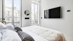 A hotel room with a bed, a TV mounted on the wall, a round mirror, and a view of the city through large windows. - Maison Labruyère - Aviau Suite (Bordeaux)