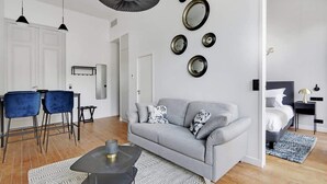 A modern living room with a grey sofa, two blue chairs, a black coffee table, and a dining area with a white table and chairs. - Maison Labruyère - Aviau Suite (Bordeaux)