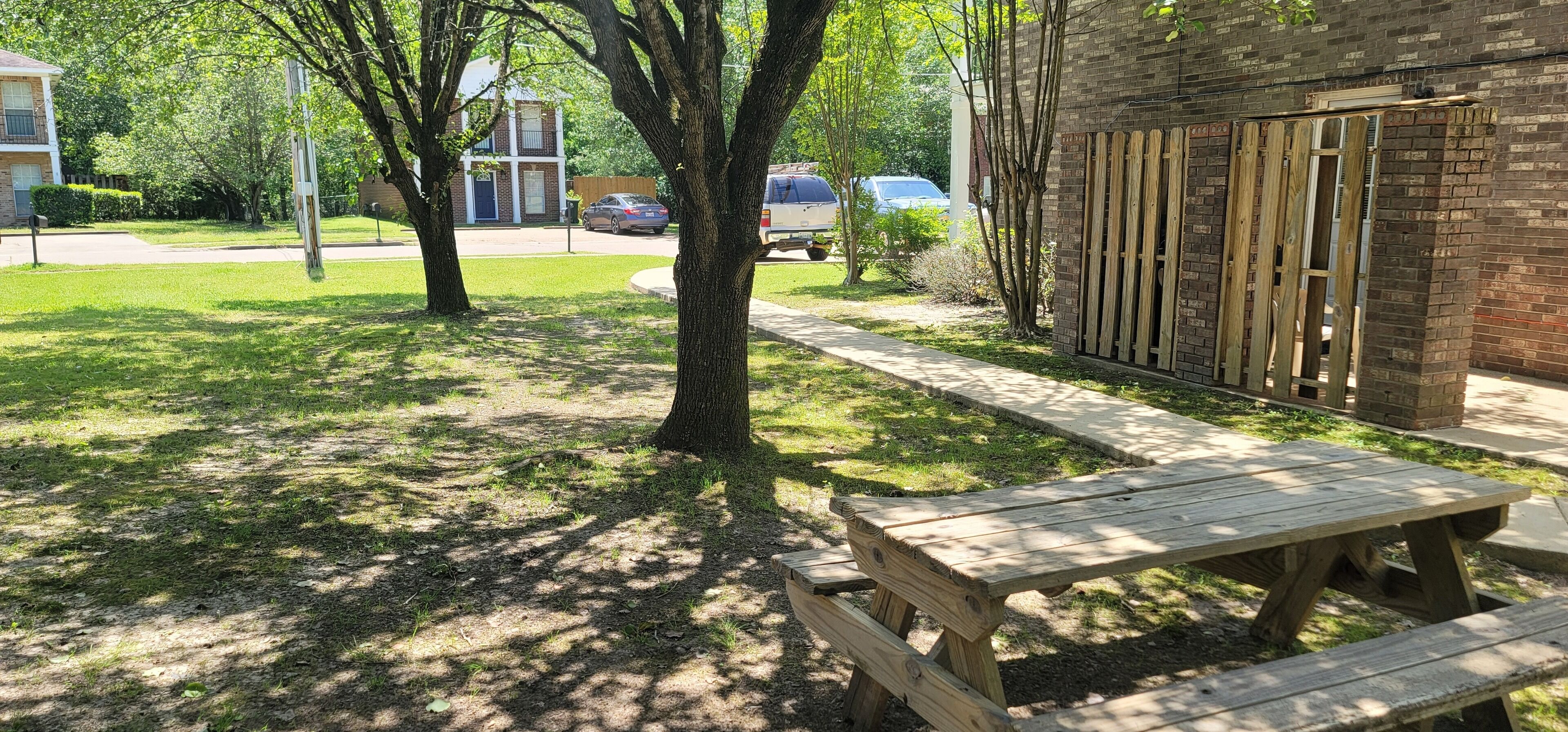 A picnic table in a grassy area with trees, a brick building, and parked cars in the background.
