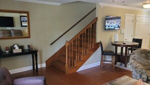 A living room with a wooden staircase, a television mounted on the wall, a dining table with chairs, and a patterned armchair. - It's Five O'Clock off South Lamar (Oxford)