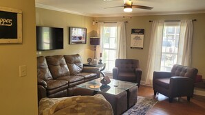 A living room with a brown leather sofa, a glass coffee table, and two armchairs. There is a flat-screen TV mounted on the wall, a framed picture, and a clock. - It's Five O'Clock off South Lamar (Oxford)