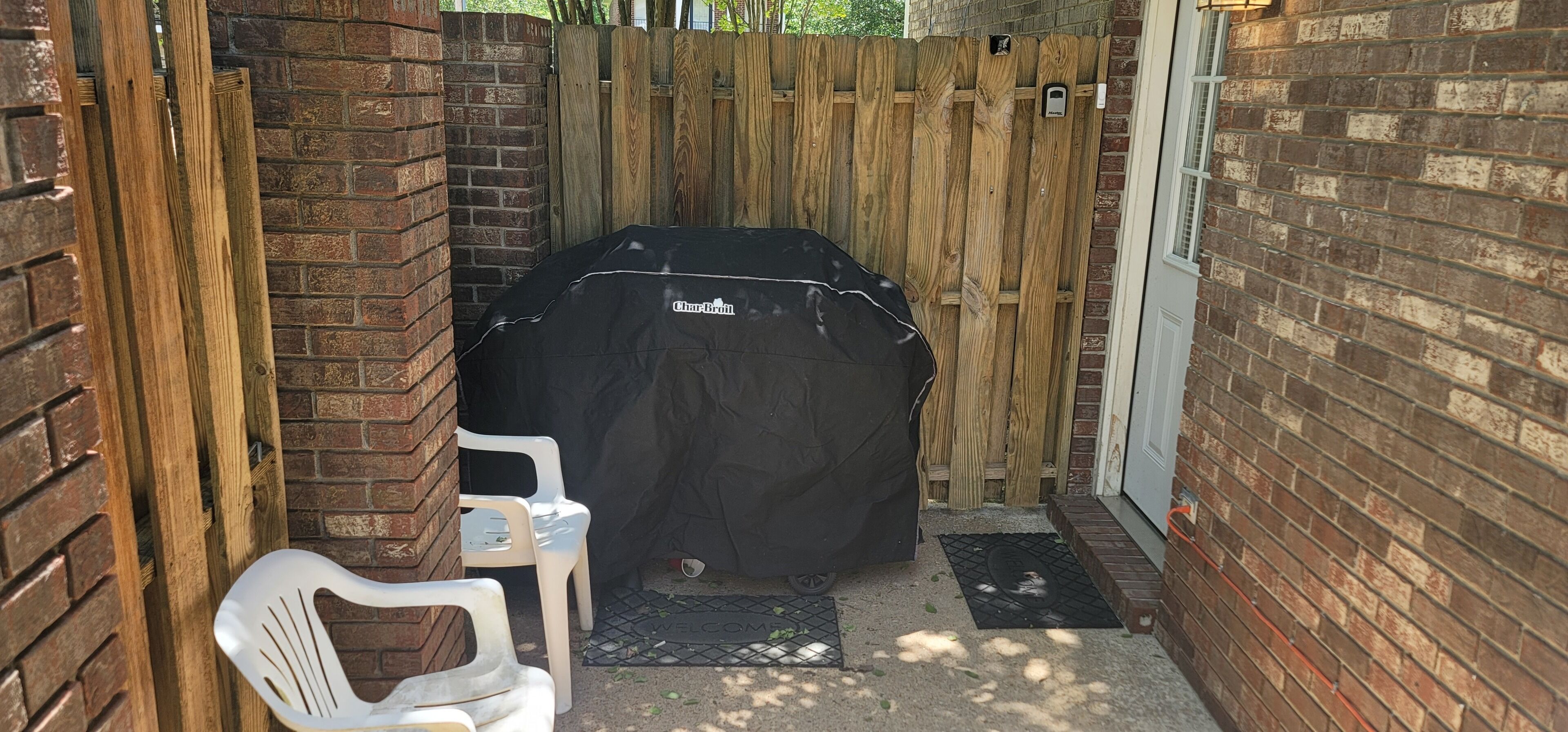 A covered barbecue grill, a white plastic chair, and a brick wall with a wooden fence in the background.