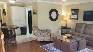 A living room with a brown leather sofa, a glass coffee table, and patterned armchairs. A dining table with chairs is visible in the background. - It's Five O'Clock off South Lamar (Oxford)