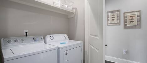 Een wasserette met twee witte voorladers wasmachines en drogers, een witte muur met twee prenten van vissen, en een wit plafond met een ronde lamp.