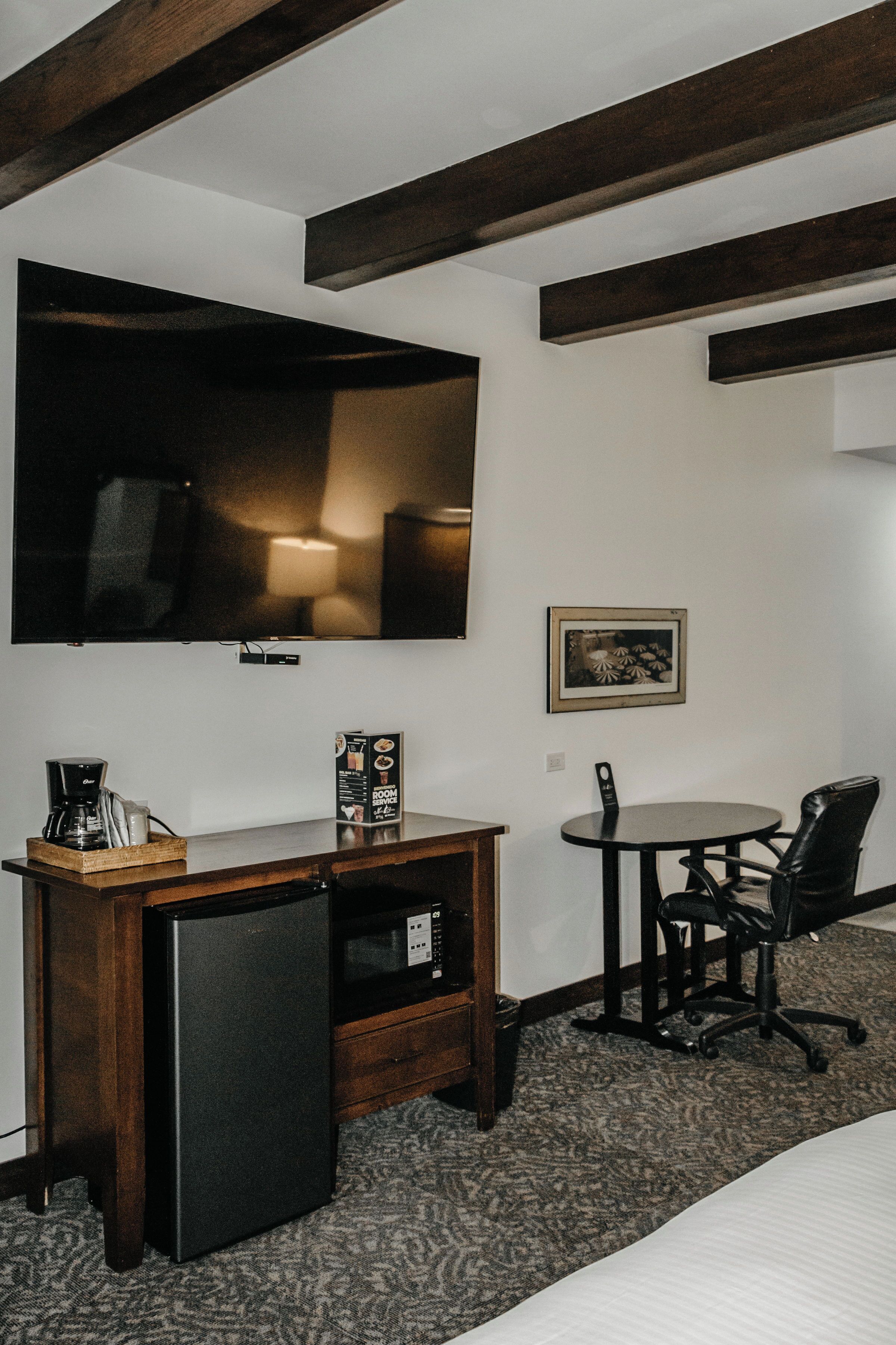 A hotel room with a flat-screen TV mounted on the wall, a small refrigerator, a coffee maker, a table with a chair, and a framed picture on the wall.