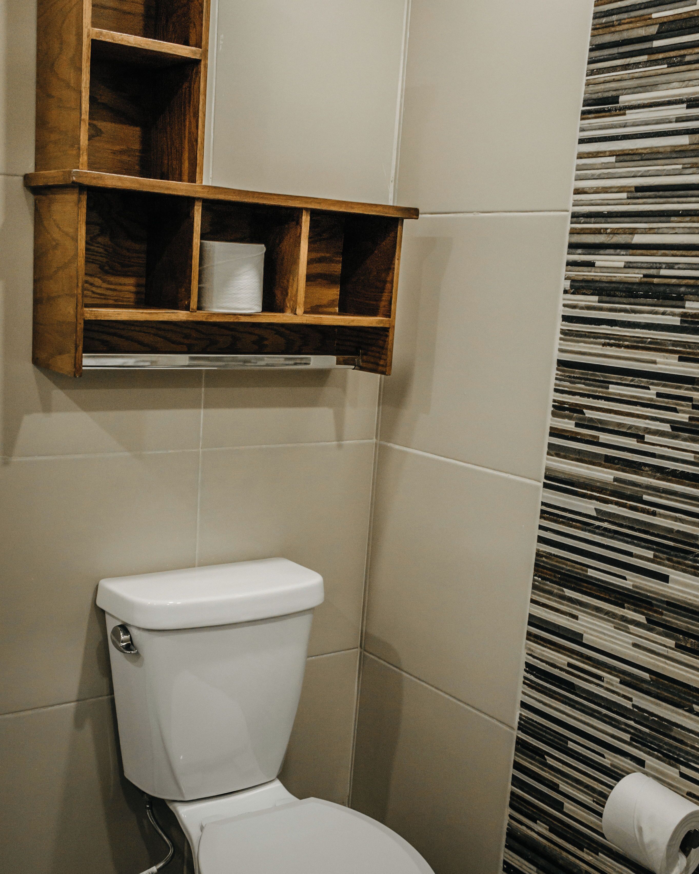 A bathroom with a toilet, a wooden shelf above it, and a patterned wall.