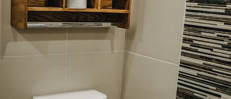 A bathroom with a toilet, a wooden shelf above it, and a patterned wall.