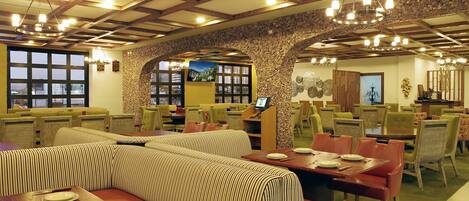 Un restaurante con techos altos, vigas de madera y una pared de piedra. Se observan varias mesas y sillas para comer, además de una amplia zona con bancos acolchados.