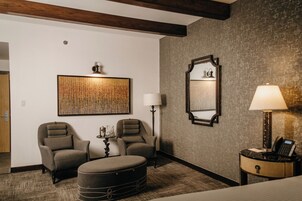 Habitación de hotel con un gran tapiz mural, dos sillones, un taburete redondo, una mesita auxiliar con lámpara, un espejo y un teléfono en una mesita de noche.
