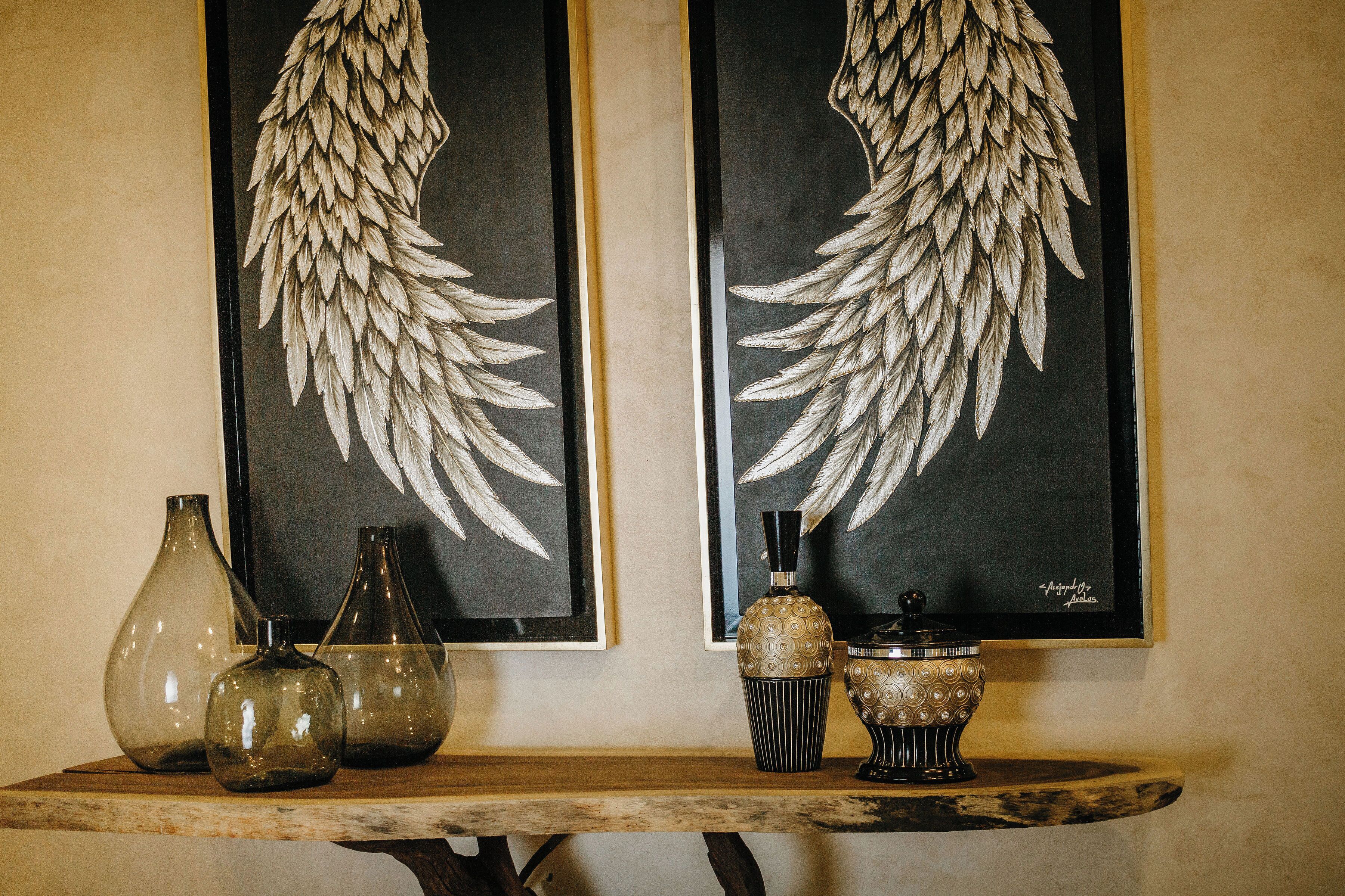 Una mesa con tres jarrones de vidrio y un cuenco decorativo, frente a una pared con dos obras de arte enmarcadas que representan alas.