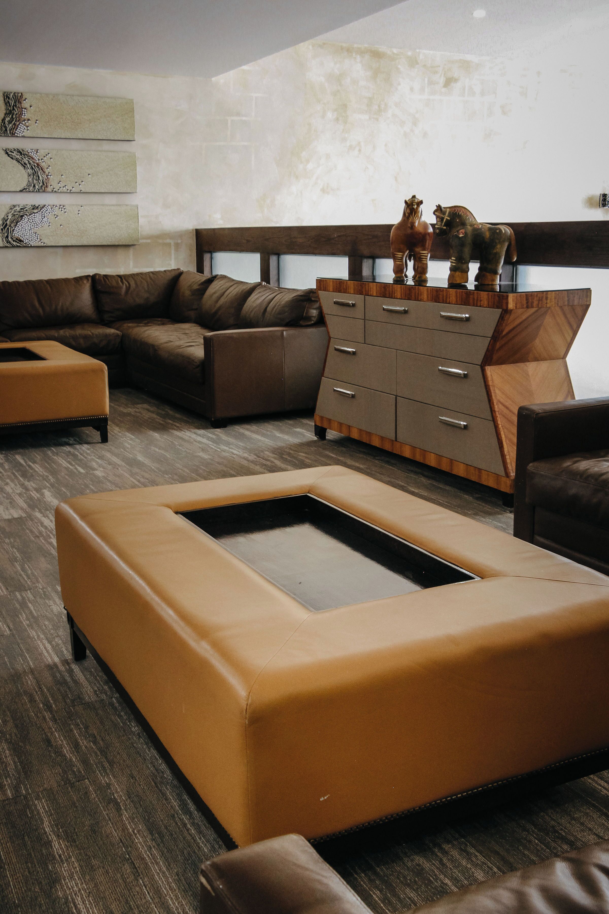 A modern living room with a brown leather sofa, a wooden cabinet with two decorative horse figures, and a large rectangular ottoman.