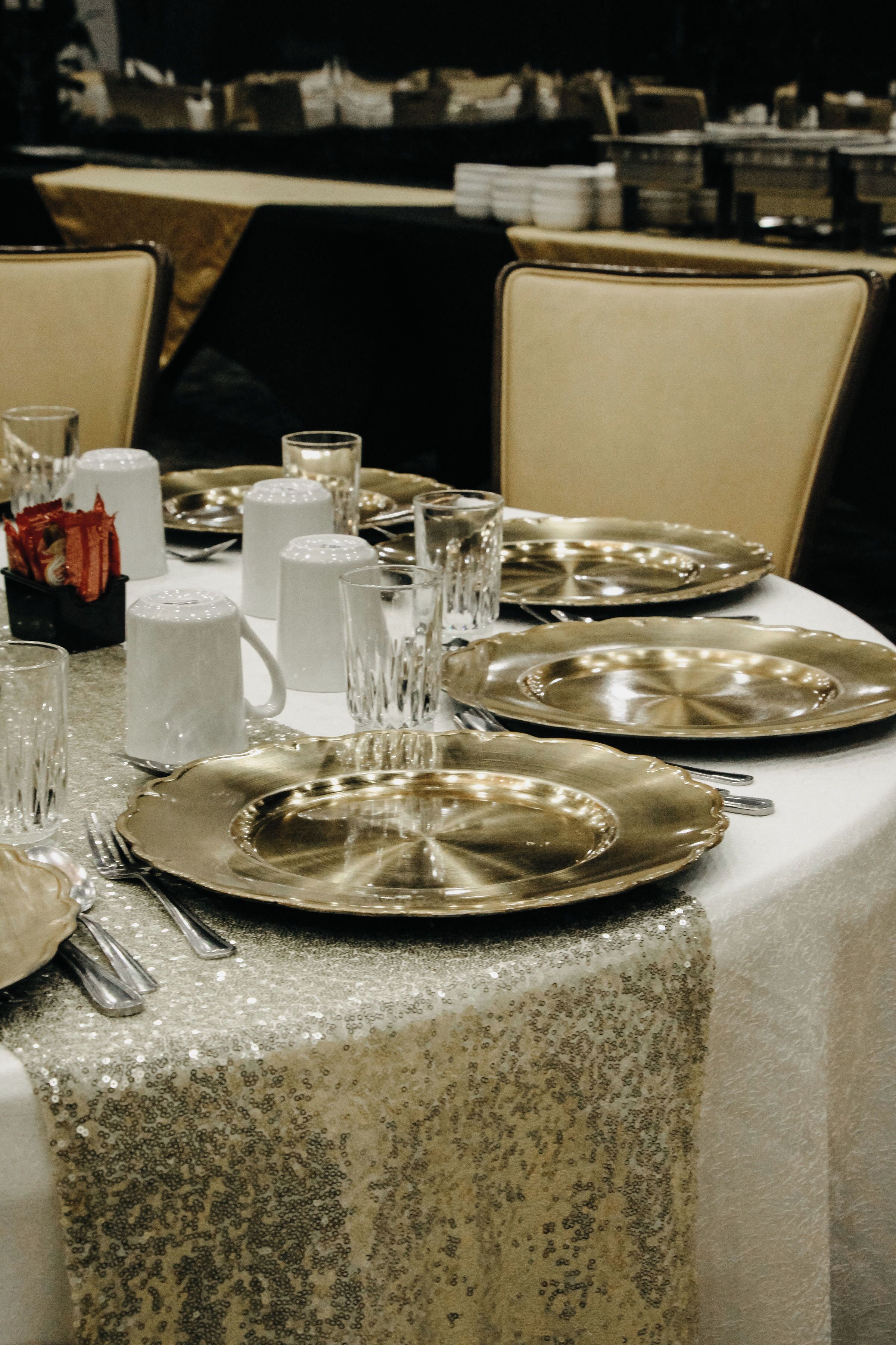 Una mesa servida con platos dorados, cubiertos de plata y vasos transparentes, cubierta con un mantel brillante.