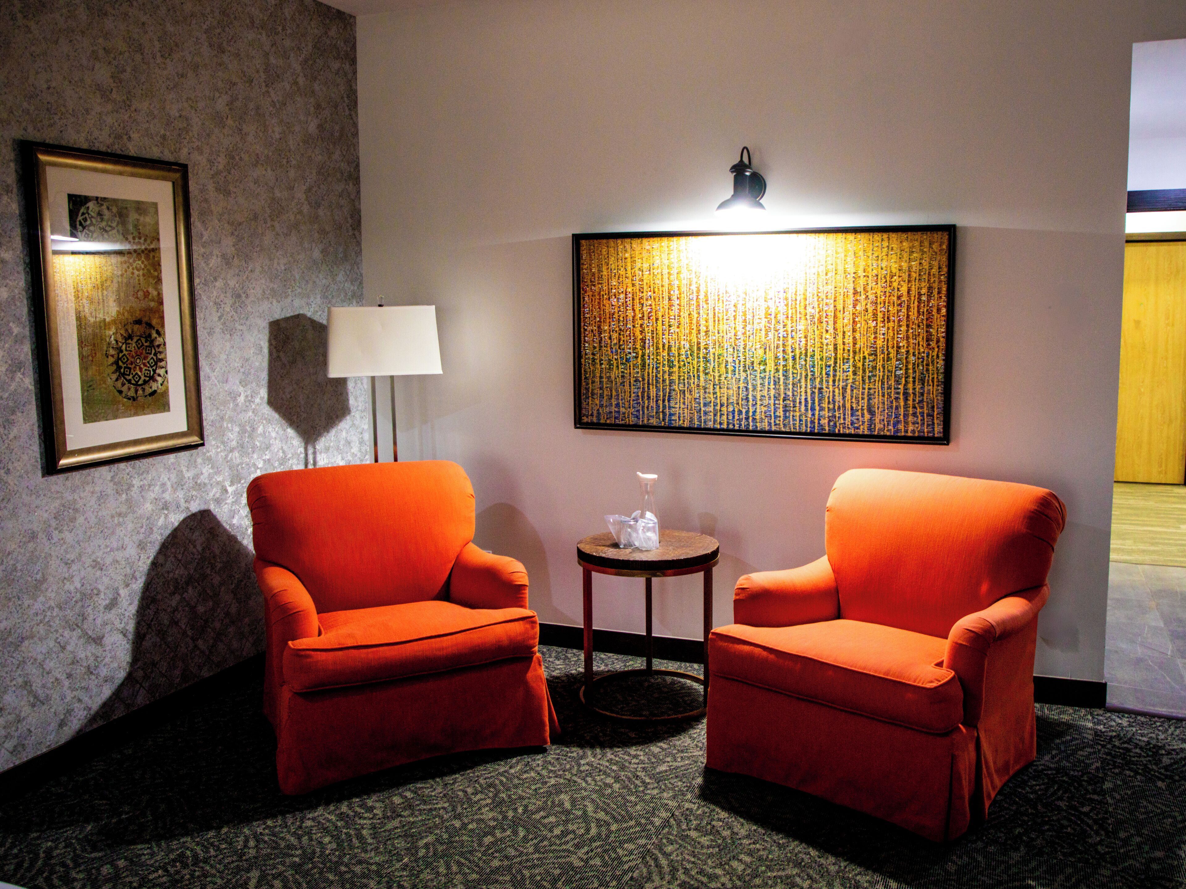 A hotel room with two orange armchairs, a small table with a lamp, a framed artwork on the wall, and a carpeted floor.