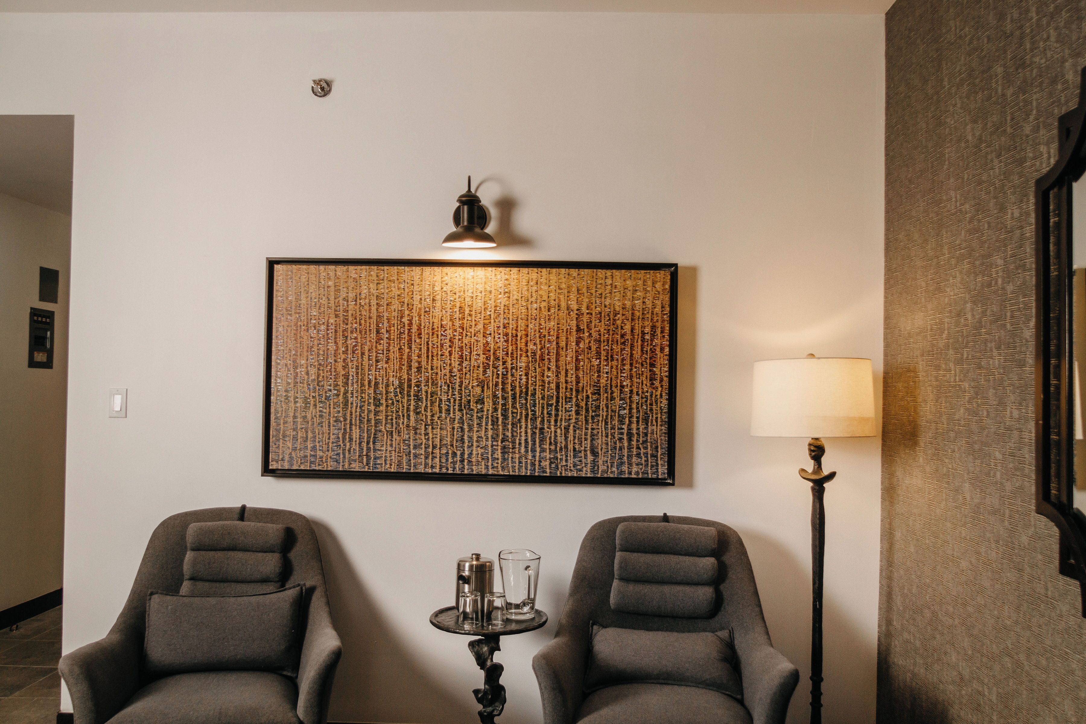 A hotel room with two grey armchairs, a small table with a lamp, and a large abstract artwork on the wall.