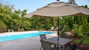 Un patio au bord de la piscine, avec une table et des chaises, un grand parasol et une piscine.