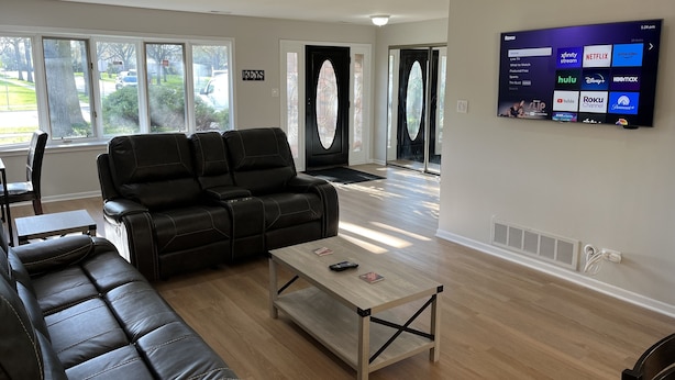 Un salon moderne avec un canapé en cuir noir, une table basse en bois et un téléviseur à écran plat fixé au mur, qui diffuse différents services de streaming.