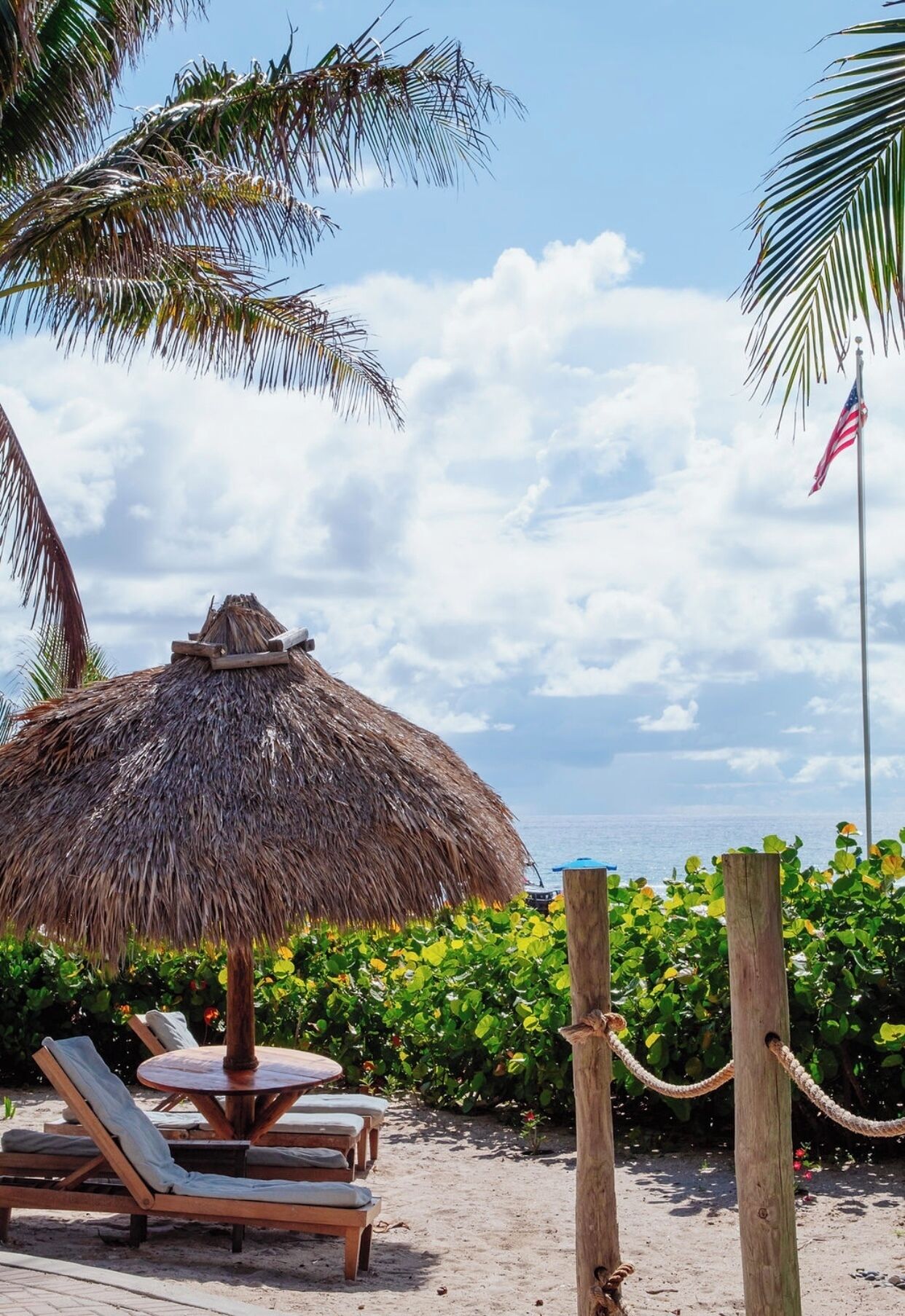 A beach with a thatched umbrella, lounge chairs, and a flag.