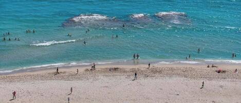 Una playa con gente nadando y tomando el sol, y varias sombrillas azules.