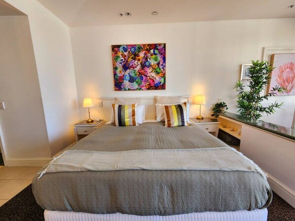 A bedroom with a large bed, two bedside tables, a nightstand, a wall-mounted painting, and a potted plant.