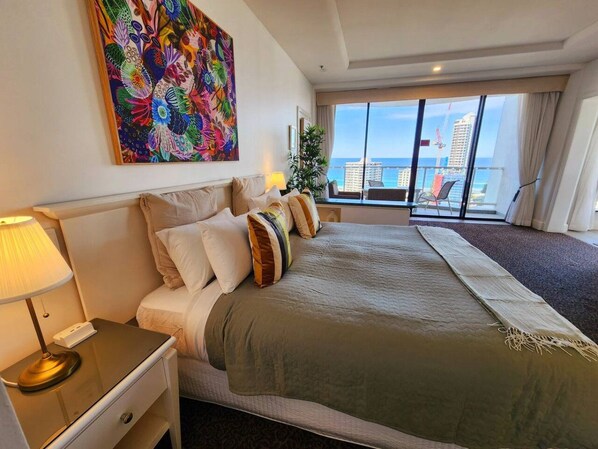 A hotel room with a large bed, a nightstand with a lamp, a painting on the wall, and a view of the city through large windows.