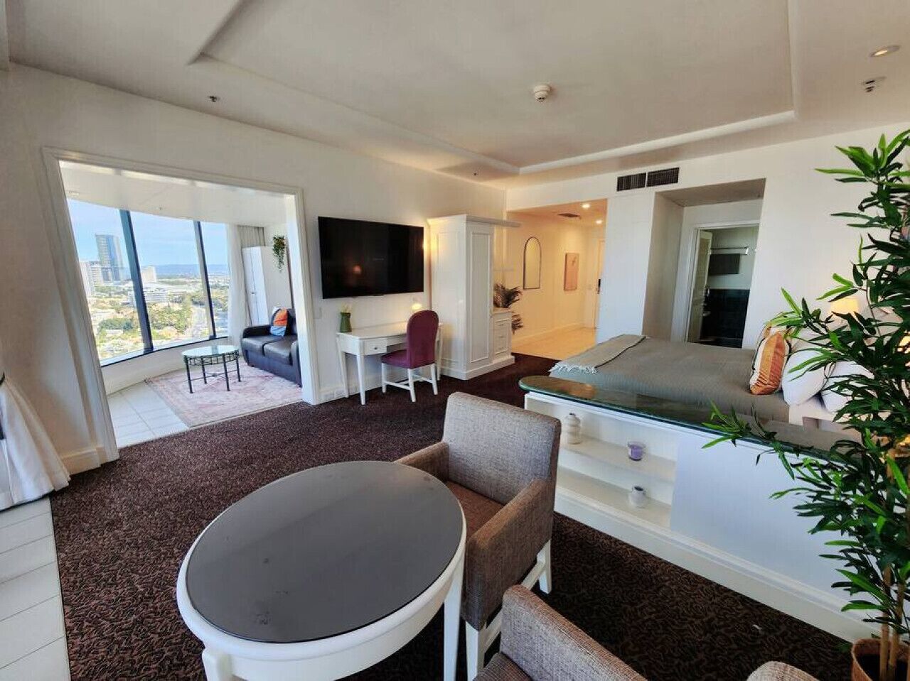 A modern hotel room with a large window, a flat-screen TV, a dining area with a round table, and a sofa.