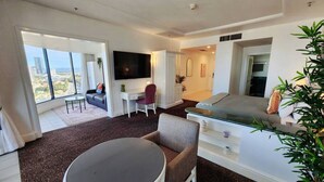 A modern hotel room with a large window, a flat-screen TV, a dining area with a round table, and a sofa.