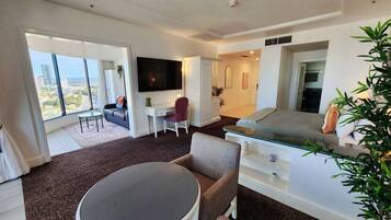 A modern hotel room with a large window, a flat-screen TV, a dining area with a round table, and a sofa.